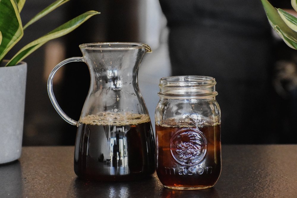 Glass carafe and mason jar filled with coffee on a dark surface.