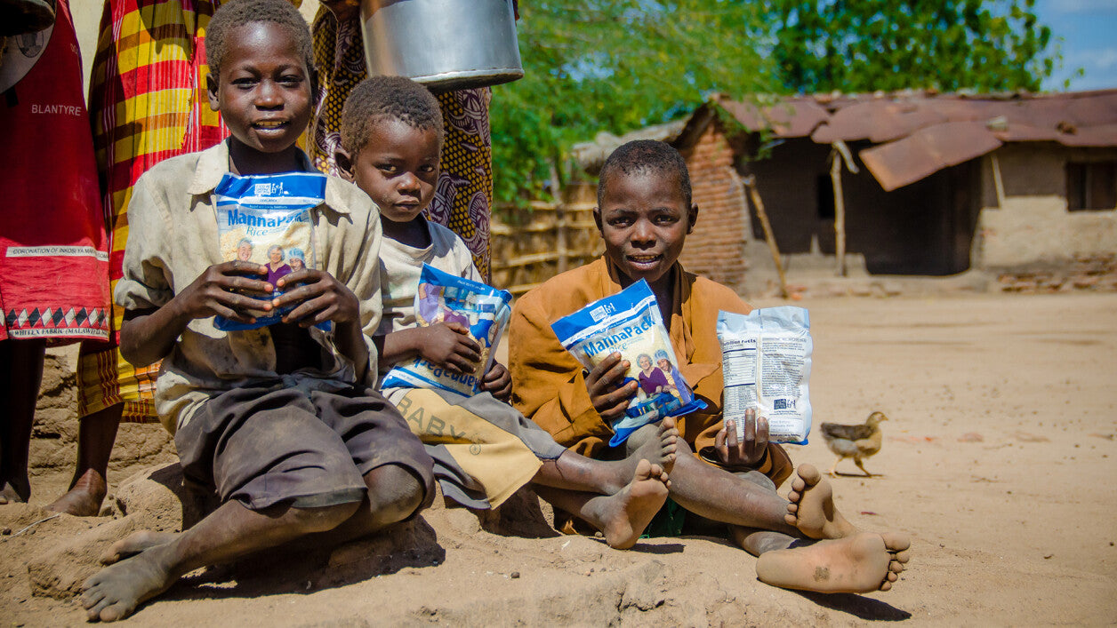Three children sitting on the ground holding packages of food in a rural setting.