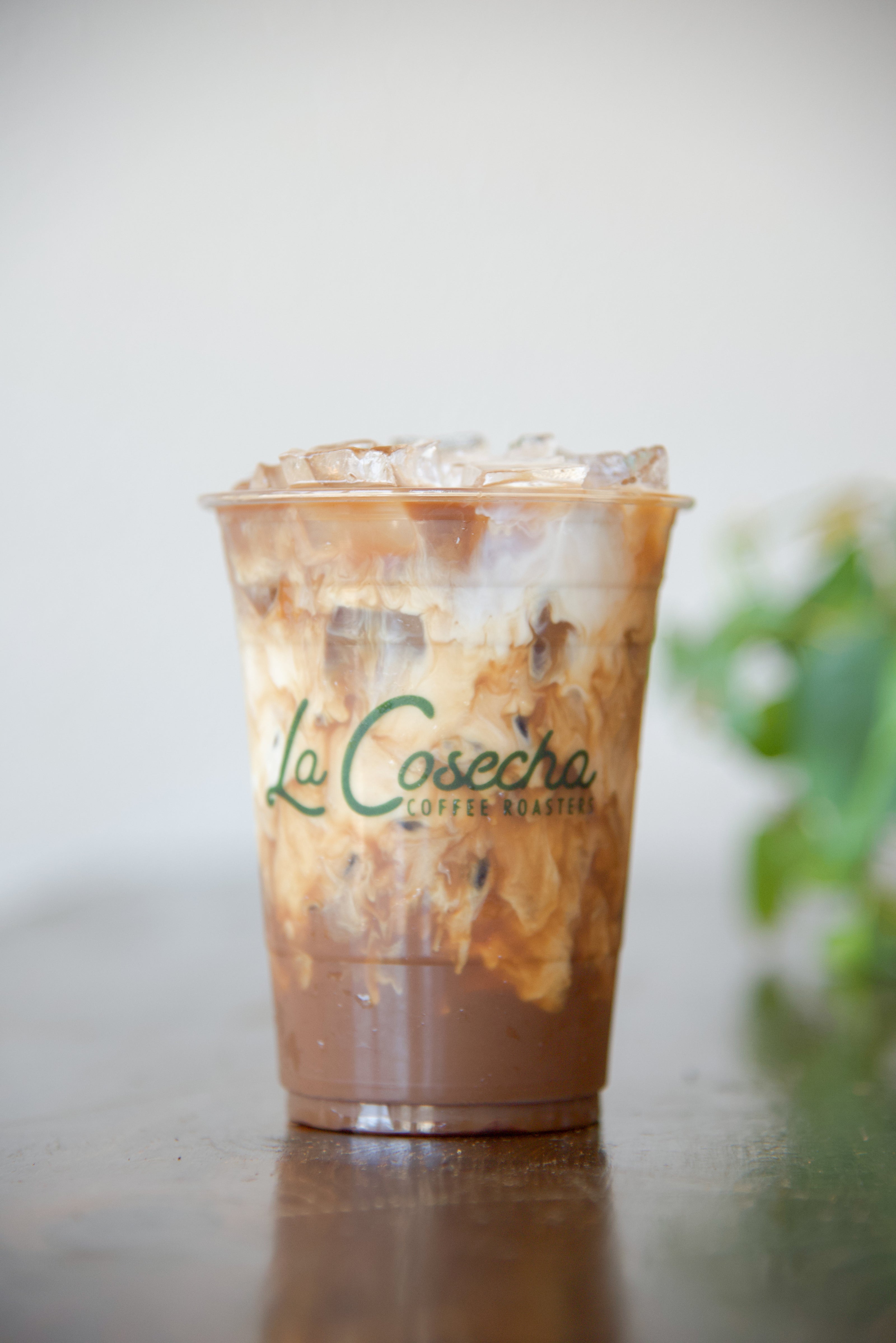 Iced coffee in a clear plastic cup with 'La Cosecha' branding on a reflective surface.
