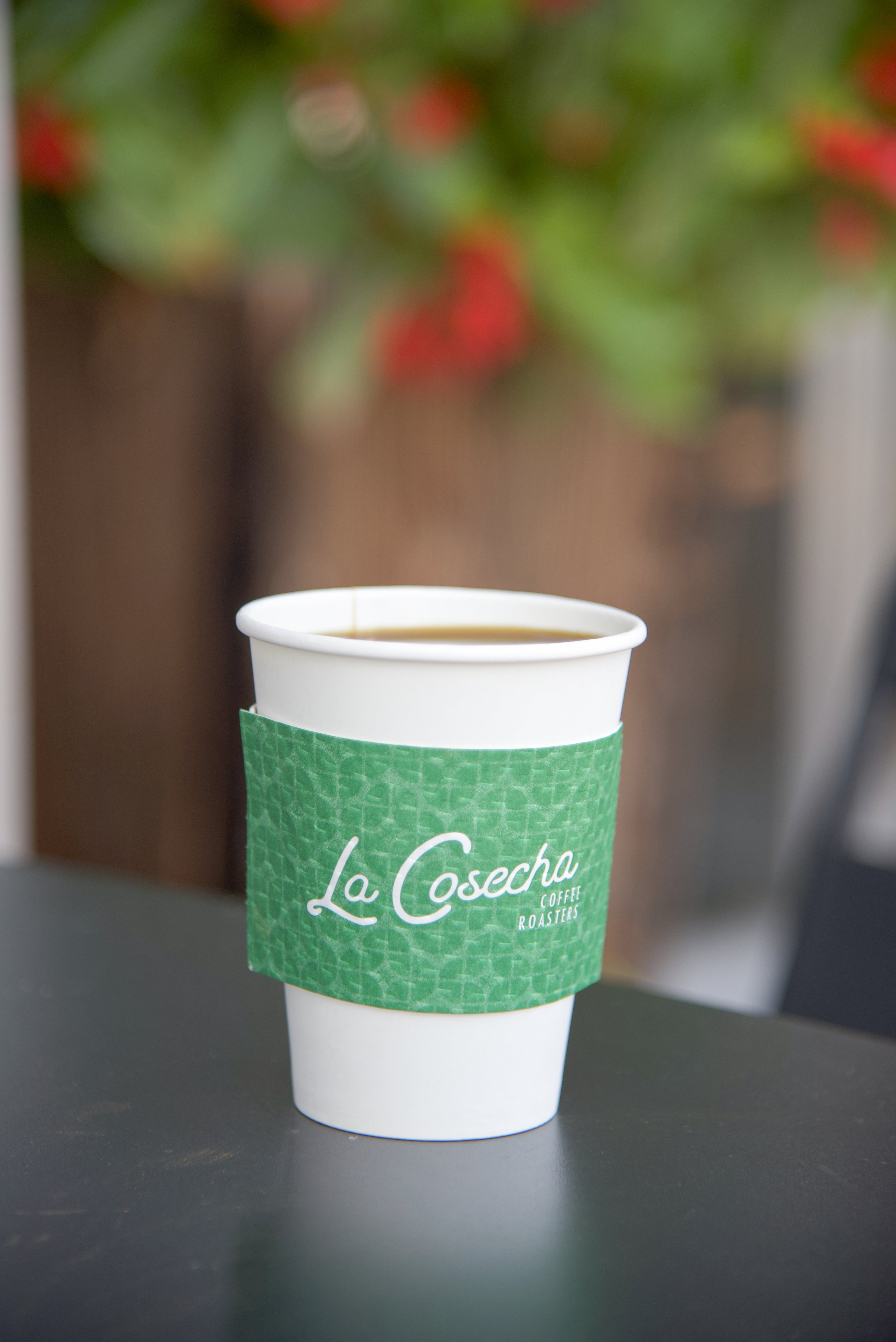 White paper cup with green sleeve featuring 'La Cosecha' on a dark surface with blurred background