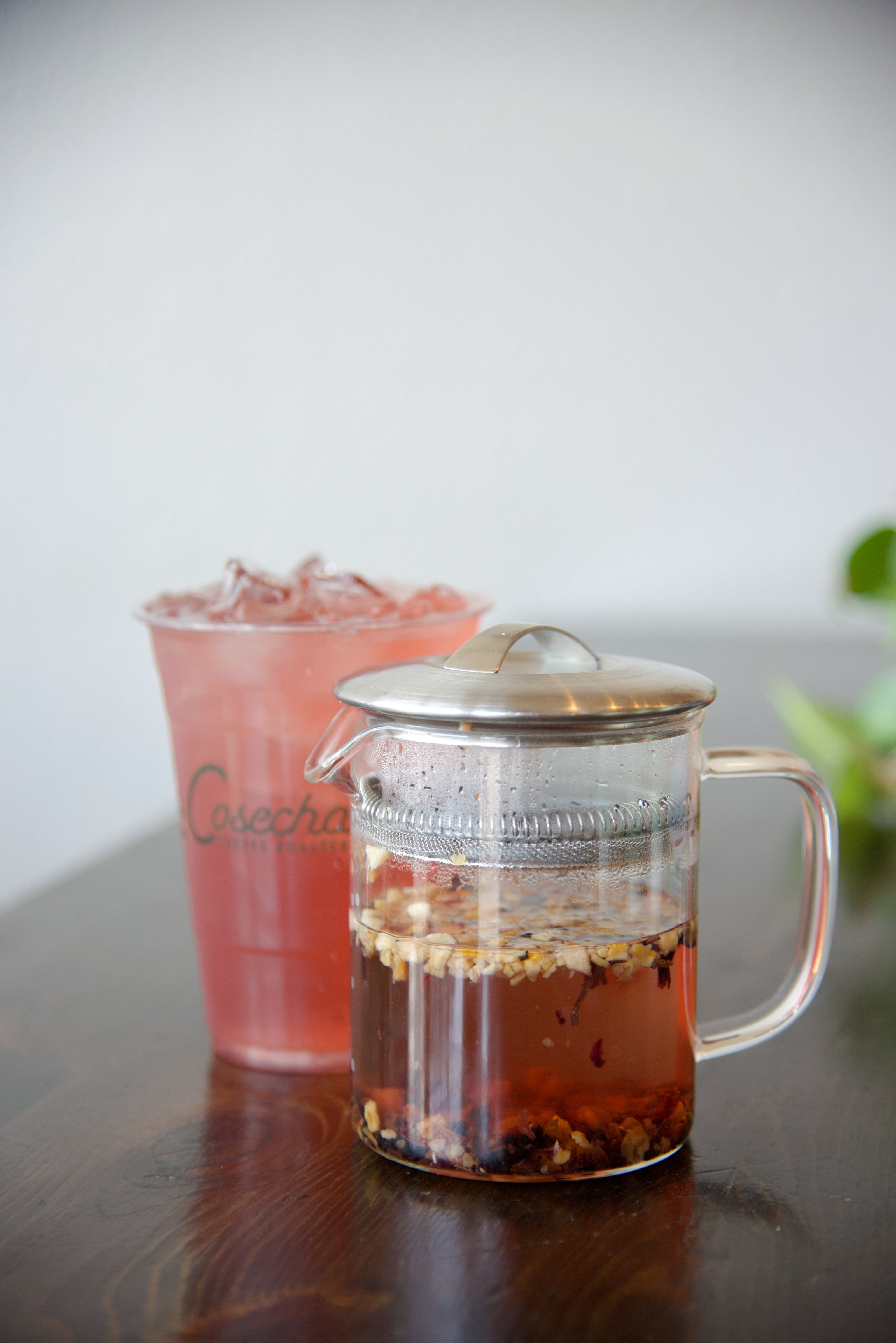 Clear glass teapot with a silver lid on a wooden surface, next to a pink drink in a glass.