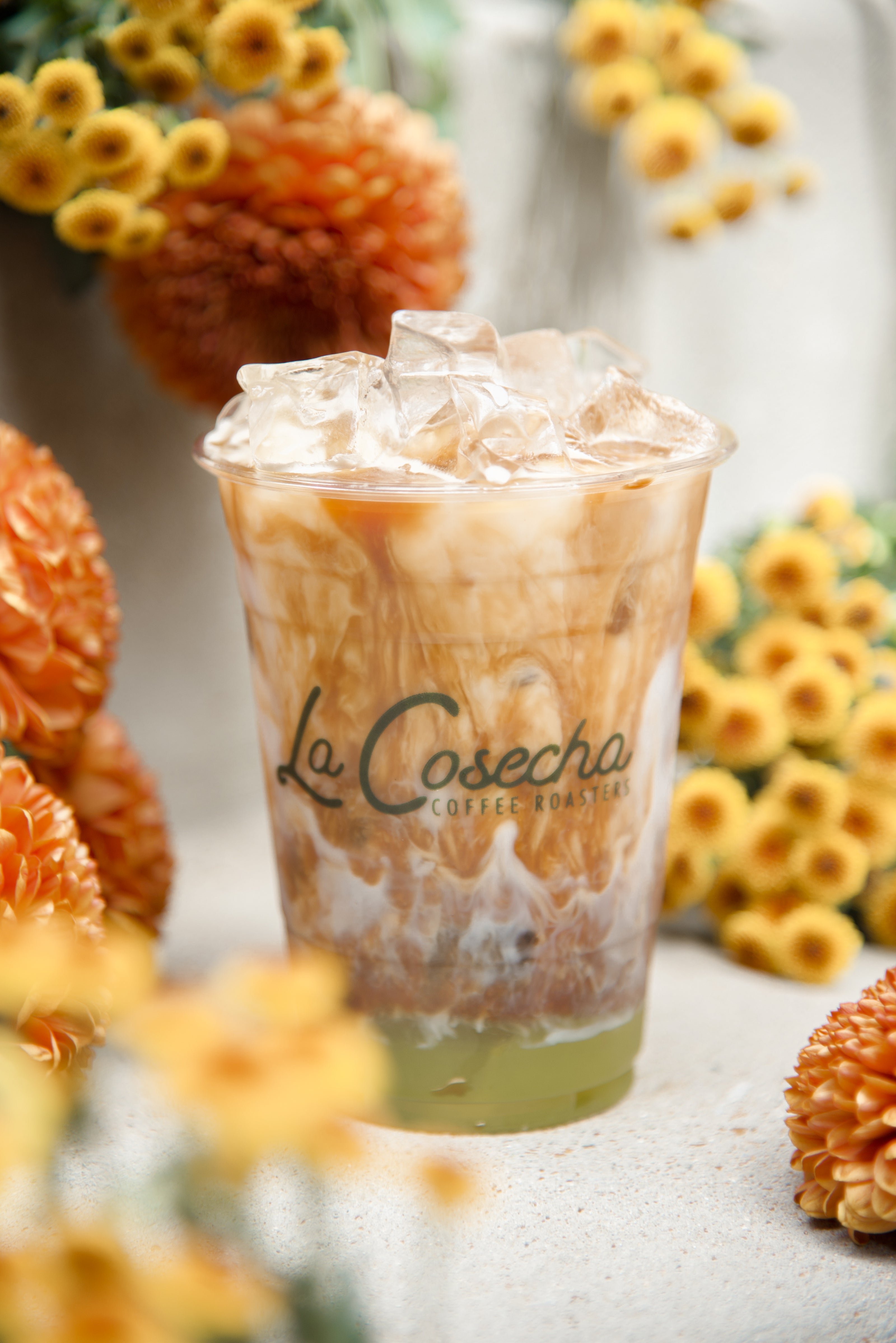 Iced coffee drink with 'La Cosecha' branding surrounded by flowers on a light surface.