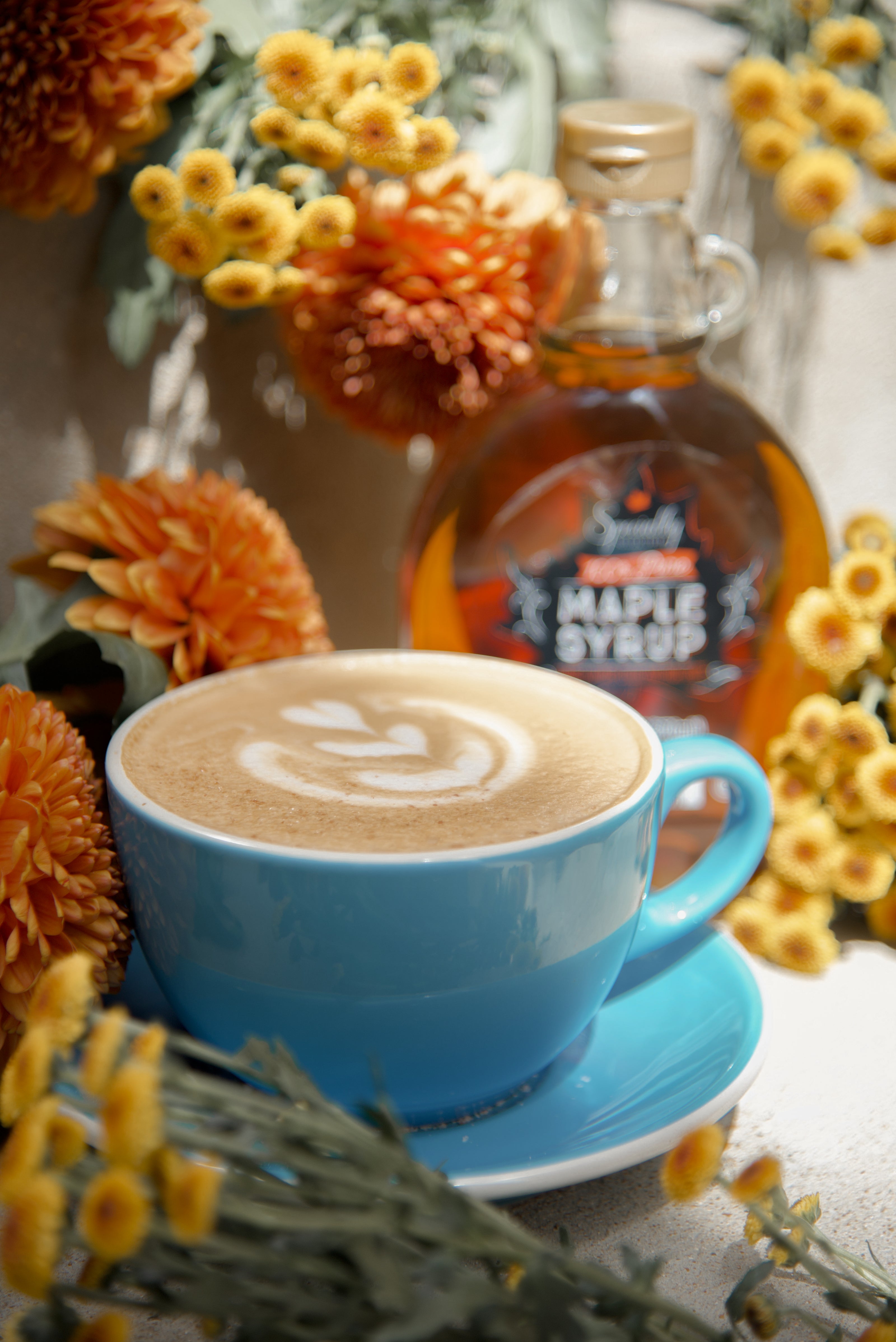 Blue cup of cappuccino with latte art, surrounded by flowers and a bottle of maple syrup.