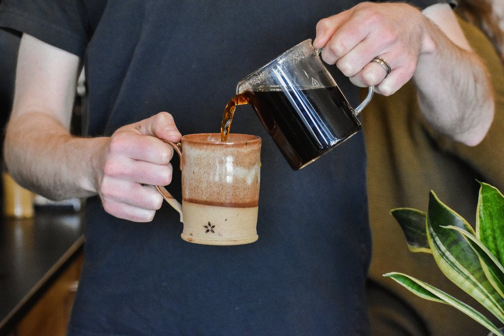 Person pouring coffee from a glass container into a ceramic mug.