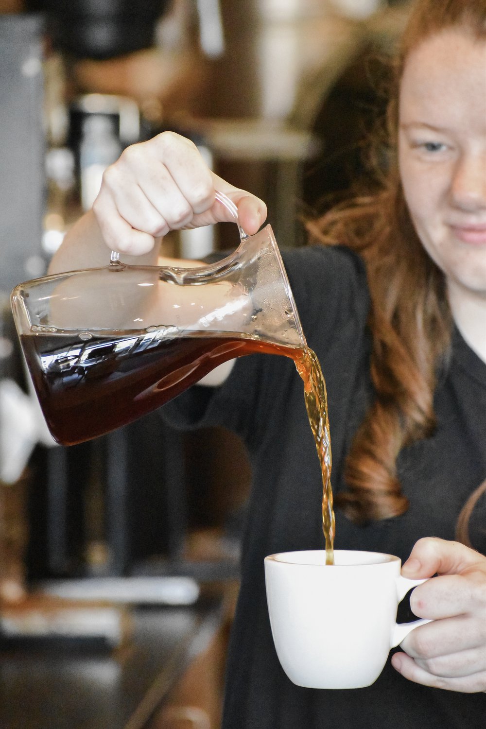 Person pouring coffee from a glass carafe into a white mug in a coffee shop.