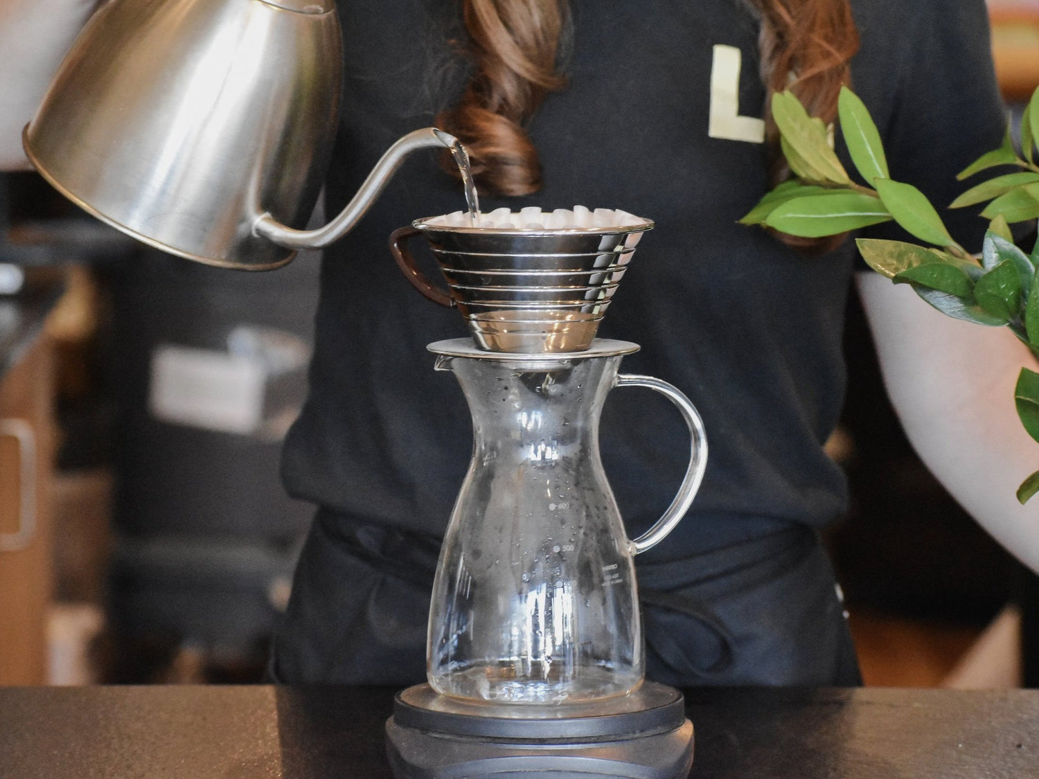 Person making coffee using a pour-over method with a clear glass carafe and metal kettle.
