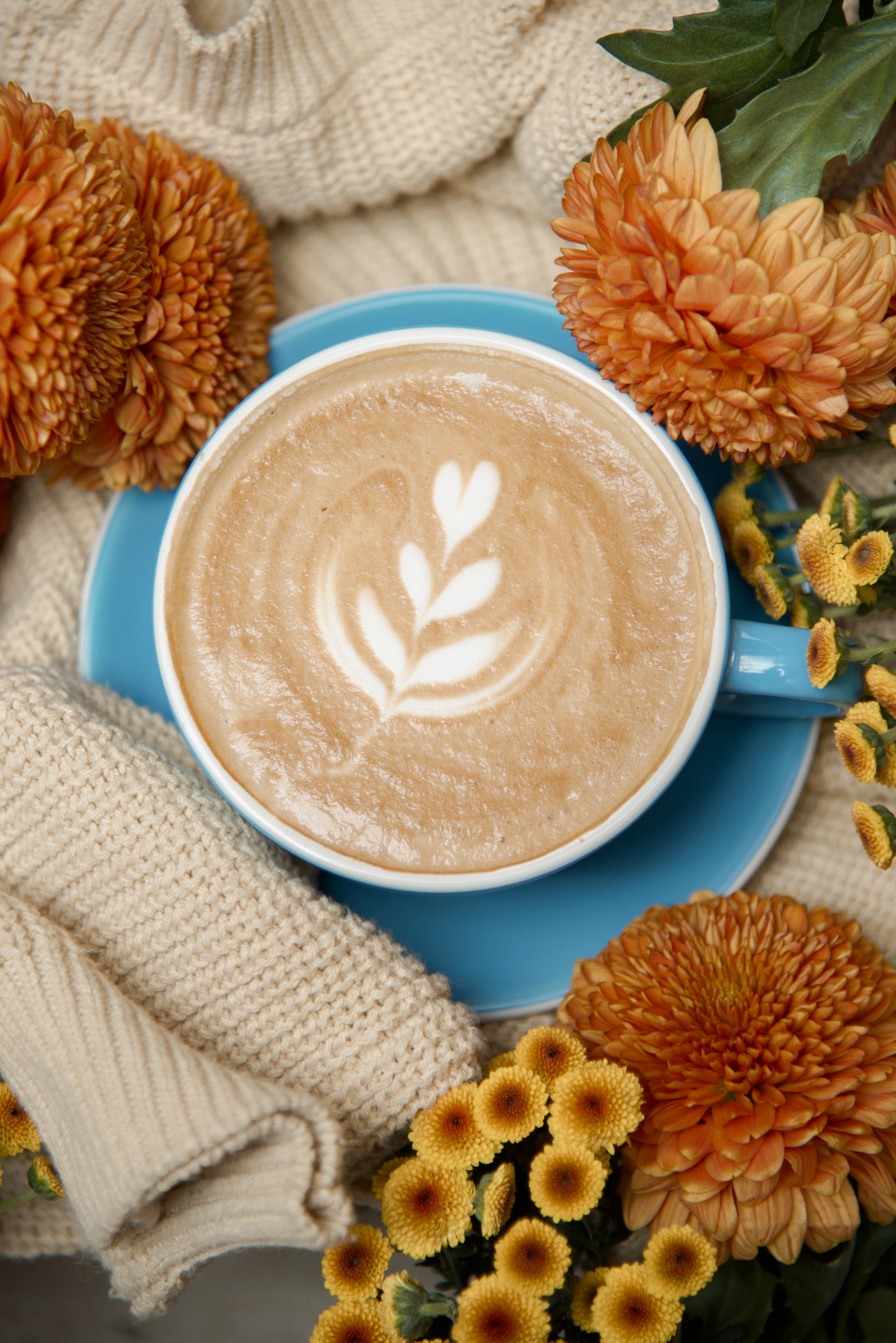 Cup of coffee with latte art on a blue saucer surrounded by autumn flowers and a scarf.